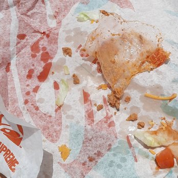 Taco Bell - Trash in your taco - The image shows a paper tray liner with remnants of a Taco Bell meal, including scattered food crumbs, small pieces of lettuce, tomato, and ground meat. A crumpled Taco Bell wrapper is partially visible in the lower left corner. The food appears to have been partially eaten, with no intact food items remaining. - Tacos Near Me - Burnet, Texas