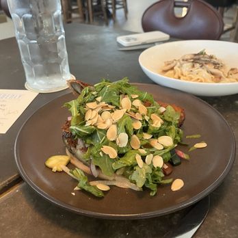 Eberly - Eggplant Milanese - A dish of eggplant Milanese with greens and sliced almonds is served on a dark plate at a restaurant table, with a glass of water and a bowl of pasta in the background. - Bars Near Me - Austin, Texas