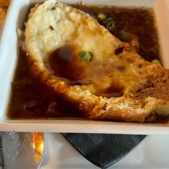 Fire Tower - French onion soup - A square ceramic bowl holds French onion soup with a layer of melted cheese on top, partially covering the dark, savory broth. Small pieces of chopped green onions are sprinkled on the surface. The bowl is placed on a white plate with a black napkin underneath, and a metal spoon rests nearby. The soup appears hot and flavorful, typical of a classic French onion dish. - New American Near Me - Stratton, Vermont