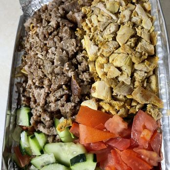 Halal Bros - The image shows a rectangular aluminum tray filled with Middle Eastern dishes. The tray is divided into sections, with ground beef on one side, chopped artichokes on another, and fresh vegetables including sliced cucumbers and diced tomatoes at the bottom. The food appears to be served in a casual setting, possibly for takeout or catering. - Middle Eastern Near Me - Austin, Texas