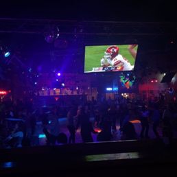 A nightclub with a dark interior illuminated by colorful lights, featuring a large screen showing a football game. The space has multiple levels, with people dancing and socializing. The environment includes a stage area, bars, and a dance floor, typical of an Austin nightlife venue.