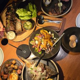 A dining table features grilled meats, a whole fish, salads, and side dishes, with utensils and small bowls, at Ossobuco in Miami.