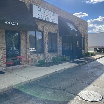 Chic Chef Catering - Chic Chef Catering - The photo depicts the storefront of Chic Chef Catering, a catering business located at 401 C-D. The building has a brick exterior with black awnings over the windows and entrance. A white sign displays the business name, and there is outdoor seating with two red chairs and a small round table. The sky is partly cloudy with sunlight shining, and the parking lot is visible in front of the building. - Caterers Near Me - Addison, Illinois
