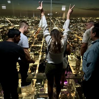 Skydeck Chicago - Five individuals are on a glass observation deck at night, overlooking a brightly lit city with streets and buildings visible below. The group includes three men and two women, with one woman in the center raising her arms and making peace signs. The deck has glass walls and a transparent floor, providing a clear view of the cityscape beneath. The scene captures a moment of sightseeing and social interaction at a high-altitude observation point. - Observatories Near Me - Chicago, Illinois