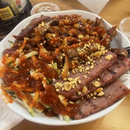 Combination Noodle Bowl's - A large white bowl filled with Vietnamese cuisine, including rice and noodle dishes topped with grilled meats, chopped nuts, and sauces. The bowl contains slices of grilled beef and pork, shredded vegetables, and is garnished with crushed peanuts and drizzled with sauces. The background shows a wooden table with condiments, drinks, and paper napkins, indicating a casual dining setting.