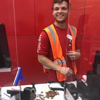 Target - Joseph helping me at the Service Desk. 5-stars! - A young man with short brown hair and a beard is smiling at the camera behind a Target checkout counter. He is wearing a red shirt with white text on the sleeve and an orange safety vest. The counter has coins, a card reader, a telephone, and a clear plastic divider. The background features red walls, indicating the store's interior. The man appears to be a Target employee assisting a customer. - Department Stores Near Me - Clearwater, Florida