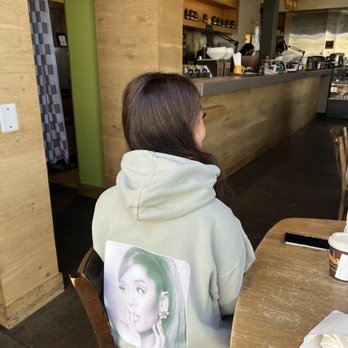 Philz Coffee - Ariana bringing her Ariana, Yo! @Philz - A woman with brown hair sits at a wooden table in a coffee shop, wearing a light-colored hoodie with a printed image of a woman on the back. The coffee shop interior features a counter with coffee-making equipment and a green wall. - Coffee & Tea Near Me - Berkeley, California
