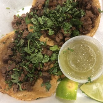 El Compa - Two tacos filled with seasoned, cooked meat and topped with chopped cilantro are presented on a plate. The tacos are accompanied by lime wedges and a small cup of green sauce, likely salsa verde. The image emphasizes traditional Mexican street food, highlighting fresh ingredients and simple presentation. - Mexican Near Me - Birmingham, Alabama