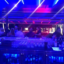 A bar scene with patrons seated and socializing under vibrant neon and LED lights, with a well-stocked bar and glasses on display.