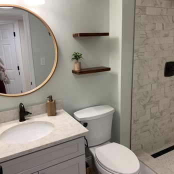 Beroli Construction - Bathroom - The image shows a bathroom with a marble vanity countertop, a black faucet, and a small potted plant. Above the vanity is a large round mirror with a wooden frame. Two wooden shelves are mounted on the wall, one with a small plant. To the right, there is a walk-in shower with marble tile walls and a black shower fixture. The bathroom has a light-colored wall and a door reflected in the mirror. - General Contractors Near Me - Southborough, Massachusetts