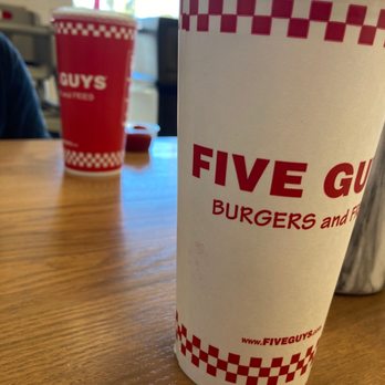 Five Guys - a cup of coffee on a table - A tall Five Guys cup with a checkered red and white design is placed on a wooden table, with another similar cup and a small container in the background. - Burgers Near Me - Mesa, Arizona