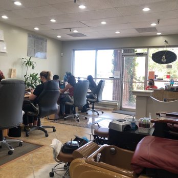 Famous Nails - The image shows the interior of a nail salon with multiple clients seated in black chairs at workstations along the wall, engaged in nail services. A large window provides natural light, and the entrance door is visible. In the foreground, there is a pedicure chair with a brown and red cushion, along with various nail and foot care tools. The ceiling has recessed lighting, and a small reception desk is near the door, with a staff member wearing a mask. The space appears clean and organized, with some plants and a menu on the wall. - Nail Salons Near Me - Albuquerque, New Mexico