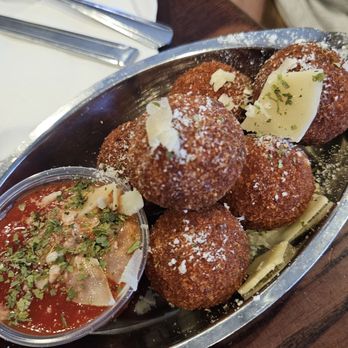 Amici Pizza - Mac & cheese arancinis - A serving of five fried arancini balls, topped with grated cheese and herbs, is presented on a metal tray. Accompanying the arancini is a small cup of marinara sauce garnished with herbs and cheese. The tray is placed on a wooden table, with a white plate, cutlery, and a person's arm visible in the background. - Pizza Near Me - Mesa, Arizona