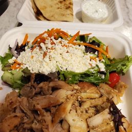 Chicken Gyro Dinner - The image shows a takeout meal with a large salad topped with crumbled cheese, shredded carrots, and mixed greens. There is grilled chicken with visible seasoning and cooking marks. A piece of pita bread is included, along with two small containers of sauces, one green and one white. The food is served in white foam containers on a marble surface.