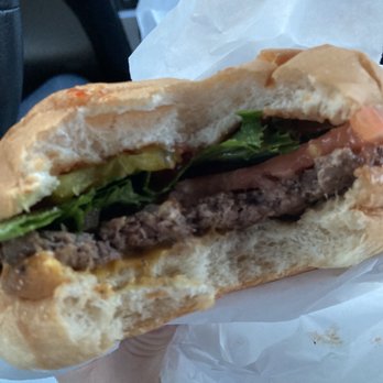 Hefner's Custard - Cheeseburger - A partially eaten cheeseburger with lettuce, tomato, and beef patty is held in a person's hand. The burger is wrapped in white paper, and the background shows the interior of a vehicle, including a seat and part of a dashboard. The bun appears soft and slightly toasted, with visible fresh vegetables and a cooked beef patty inside. - Ice Cream & Frozen Yogurt Near Me - Cedarburg, Wisconsin