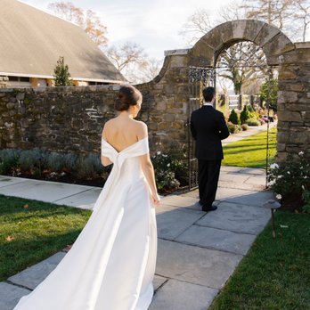 L'elite Bridal - A bride in a strapless white wedding gown with a long train walks along a stone pathway toward a groom standing near a stone archway. The groom is dressed in a black suit and is facing away from the camera. The setting is outdoors with a garden, trees, and a building with a sloped roof in the background, illuminated by natural sunlight. - Bridal Near Me - Boston, Massachusetts