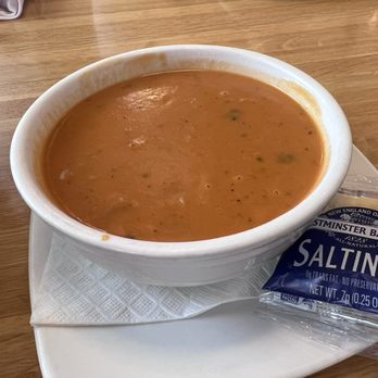 Silver Diner - Tomato basil soup - A ceramic bowl filled with tomato basil soup is positioned on a white plate, with a packet of saltines crackers placed next to it. The table surface is wooden, and the soup appears smooth with visible herbs. The setting suggests a casual dining environment, possibly at a restaurant or cafe. - Diners Near Me - McLean, Virginia