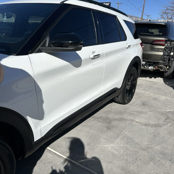 Body Master - Car - The image shows a white SUV with black wheels and tinted windows parked on a concrete surface outdoors. The vehicle's side mirror is black, and the door handles are white. In the background, there is another vehicle with visible damage to its rear, and utility poles with power lines are also visible against a clear blue sky. The shadow of the person taking the photo is cast on the ground. - Body Shops Near Me - Albuquerque, New Mexico