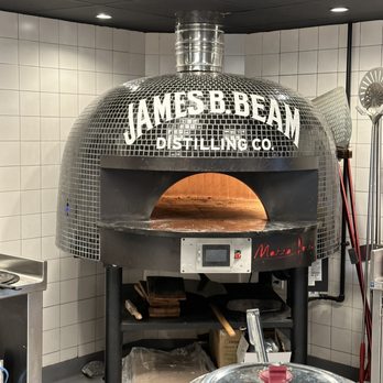 The Kitchen Table - Gas fired pizza oven. - A large, dome-shaped pizza oven with a mosaic tile exterior displaying the branding 'James B. Beam Distilling Co.' on the front. The oven has a metallic chimney on top and is situated in a commercial kitchen with white tiled walls and ceiling lights. There are kitchen tools and equipment nearby, indicating a food preparation environment. - American Near Me - Clermont, Kentucky