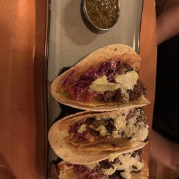 Tommy Gun Tacos - Three tacos are arranged on a rectangular plate, each filled with different ingredients including meat, shredded vegetables, and sauces. The tacos are topped with cheese and other garnishes. A small metal bowl containing a green salsa or sauce is placed above the tacos. The plate is set on a wooden table, and part of a person's leg is visible in the background.