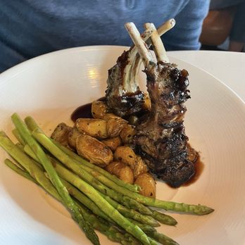 Holman's Table - Veal Rack - A white plate holds two grilled veal chops with bones, glazed with a dark sauce. Accompanying the meat are roasted potatoes and steamed asparagus spears. The presentation emphasizes the meat as the main focus, with the vegetables and potatoes arranged around it. The background shows a person wearing a blue shirt, indicating a dining setting. - Bars Near Me - Saint Paul, Minnesota