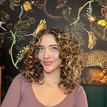 Salon Estique - So happy with my results by Emma - A woman with shoulder-length, curly blonde hair and blue eyes is seated indoors against a mural featuring jungle animals and plants. - Hair Salons Near Me - Phoenix, Arizona