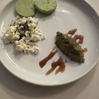 Joon - The image shows a white round plate with three food items: two slices of green hummus, crumbled white cheese with black seasoning, and a meatball topped with a drizzle of sauce. The plate is placed on a white surface, and the food presentation is simple and minimal. - Persian/Iranian Near Me - Vienna, Virginia