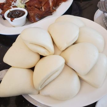 Tasty Kitchen - A white plate holds multiple steamed buns, arranged in a pile, with a smooth, rounded appearance. In the background, there is a dish of roasted meat with a dark sauce on the side, garnished with shredded vegetables. The setting appears to be a restaurant table with other dishes partially visible. - Cantonese Near Me - Davis, California