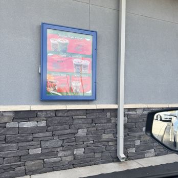 Dutch Bros Coffee - A framed menu is mounted on a gray exterior wall, showing various coffee drinks and prices, with a stone lower wall and a parking lot visible. - Coffee & Tea Near Me - Albuquerque, New Mexico