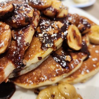 Elly's Pancake House - Stacked pancakes with sliced bananas, drizzled with chocolate syrup, and sprinkled with powdered sugar, served on a white plate. - Breakfast & Brunch Near Me - Norridge, Illinois
