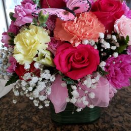 A floral bouquet featuring roses, carnations, baby's breath, and a pink butterfly ornament, arranged in a green container with pink ribbon, on a granite surface.
