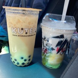 Good milk tea.   Customer service is ok - Two bubble tea drinks are positioned on a car dashboard. The left drink is a large cup with a clear lid, containing a light-colored beverage with black tapioca pearls at the bottom and a black straw. The cup has the logo 'KUTEA' and a website URL. The right drink is a smaller cup with a clear lid, containing a layered beverage with white, black, and green elements, and a white straw. The background shows car seats and a person wearing a blue jacket, indicating the photo was taken inside a vehicle.