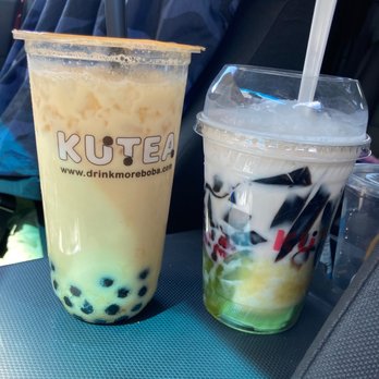 KUTEA - Good milk tea.   Customer service is ok - Two bubble tea drinks are positioned on a car dashboard. The left drink is a large cup with a clear lid, containing a light-colored beverage with black tapioca pearls at the bottom and a black straw. The cup has the logo 'KUTEA' and a website URL. The right drink is a smaller cup with a clear lid, containing a layered beverage with white, black, and green elements, and a white straw. The background shows car seats and a person wearing a blue jacket, indicating the photo was taken inside a vehicle. - Desserts Near Me - Denver, Colorado