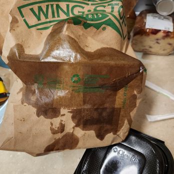 Wingstop - Sauce leaked all out these cheap containers. - A Wingstop paper bag with visible sauce stains is positioned on a table, with a black plastic food container nearby. The background includes other food items and containers, indicating a meal or takeout setting. - Chicken Wings Near Me - Laurel, Maryland