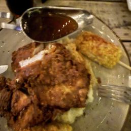 Yard Bird - A plate contains fried chicken pieces, two corn on skewers, and a small cup of dark sauce. The food is placed on a glass plate, and the background shows a wooden table surface and part of a restaurant interior. The image is slightly blurry and taken from a top angle.