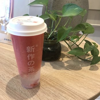 Sun Tea - Peach strawberry tea slush with agar and jelly. #4 bestseller - A pink and white layered drink in a tall plastic cup with a white lid is positioned on a wooden table. The drink appears to be a fruit tea or slush, with visible ice or jelly at the bottom. Behind the cup, there is a black ceramic pot containing a green leafy plant, and a decorative stone object with a circular cutout. The setting suggests a casual cafe or tea shop environment. - Bubble Tea Near Me - Markham, Ontario