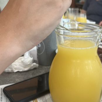 Another Broken Egg Cafe - Pitcher of Mimosa - A person’s hand with a ring is stirring a pitcher of orange juice on a restaurant table, with a smartphone and a menu visible. - Breakfast & Brunch Near Me - Fishers, Indiana