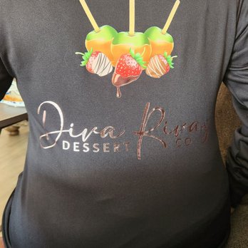 Epic Imprints - Custom logo jacket - The image shows a person seated, wearing a black jacket with a printed logo on the back. The logo depicts three caramel apples with sticks and strawberries dipped in chocolate, with the text 'Diva Riva Dessert Co.' below the graphic. The setting appears to be indoors, with some decorations visible in the background. - Embroidery & Crochet Near Me - Baldwin Park, California