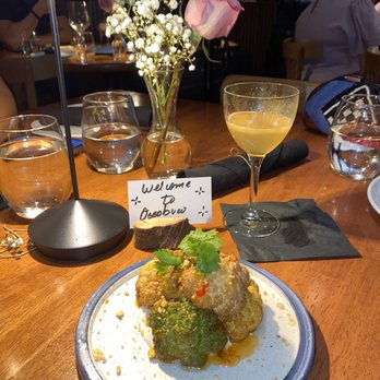 Ossobuco Miami- Steak house - SWEET & SOUR CAULIFLOWER - Fried cauliflower with herbs and sauce on a white plate, with a glass of yellow cocktail, flowers, and glasses of water on a wooden table at Ossobuco. - Steakhouses Near Me - Miami, Florida
