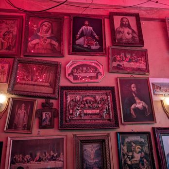 The Thin Man - The image shows a wall adorned with multiple framed religious paintings and photographs, including images of Jesus Christ, the Last Supper, and other religious figures. The wall is illuminated by pink lighting, with visible electrical wires hanging from the ceiling. The artwork varies in size and style, with some pieces in ornate frames and others in simpler borders, creating a collage-like display. - Bars Near Me - Denver, Colorado