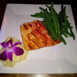 Grilled Salmon - A square white plate contains a piece of grilled salmon, a serving of green beans, and a purple flower with a yellow garnish, arranged as a meal presentation.