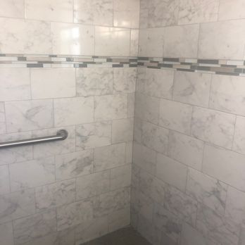 Budget Inn - marble print tile - Shower area with marble tile walls, a horizontal decorative tile strip, metal grab bars, and a hexagonal tile floor with a drain. - Hotels Near Me - Saint Augustine, Florida