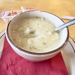 Cup of Cream of Chicken w/Green Chile Soup - A ceramic bowl filled with cream of chicken with green chile soup sits on a red paper napkin on a wooden table. The soup appears creamy with visible green herbs and chunks of chicken or vegetables. A metal spoon is partially submerged in the soup. In the background, there are some packaged bread or crackers and part of a person's arm is visible at the top of the image.