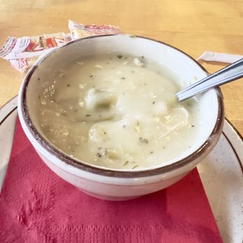 Christy Mae's Restaurant - Cup of Cream of Chicken w/Green Chile Soup - A ceramic bowl filled with cream of chicken with green chile soup sits on a red paper napkin on a wooden table. The soup appears creamy with visible green herbs and chunks of chicken or vegetables. A metal spoon is partially submerged in the soup. In the background, there are some packaged bread or crackers and part of a person's arm is visible at the top of the image. - Sandwiches Near Me - Albuquerque, New Mexico