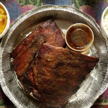 Mr E Chefs Barbecue - A meal featuring barbecue ribs with a dark, glazed exterior, served on a round metal tray. The tray includes a small container of barbecue sauce. Two side dishes are present: a bowl of potato casserole with melted cheese and a bowl of coleslaw with shredded vegetables. The tray is placed on a colorful, patterned tablecloth. - Caterers Near Me - Chicago, Illinois