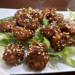 Double Fried Chicken - A plate of Korean double fried chicken features crispy, glazed chicken pieces topped with sesame seeds, arranged on a bed of shredded lettuce. The chicken appears to be coated in a thick, flavorful sauce, highlighting Korean culinary influences. The dish is presented on a white plate, emphasizing the contrast between the dark sauce and the fresh green lettuce.