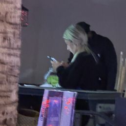 Evidence - A woman with blonde hair is seated at a table, focused on her phone, with a man standing behind her in a dark environment.