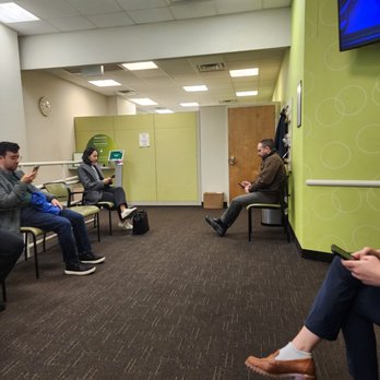 Quest Diagnostics - The image depicts a waiting room with five people seated on chairs, each using their smartphones. The room has a green accent wall, a wooden door, and a wall-mounted television. The ceiling features fluorescent lighting and air vents. The floor is carpeted in dark brown, and there is a clock on the wall. The setting appears to be a medical or office waiting area. - Laboratory Testing Near Me - Brooklyn, New York