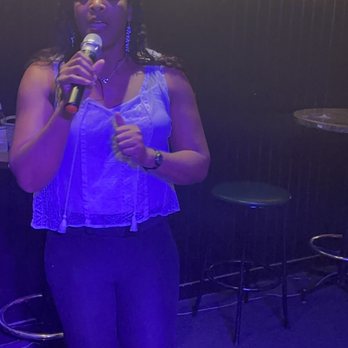 Kip's Pub - One amazing singer after another - A woman is standing and singing into a microphone in a dimly lit venue, possibly a bar or karaoke space. She is wearing a sleeveless top, dark pants, and earrings, with her eyes closed and a focused expression. The background features a chalkboard menu with colorful writing and a black wall with a strip of yellow lighting. There are bar stools and small tables visible behind her. - Pool Halls Near Me - Indianapolis, Indiana