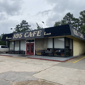 105 Cafe - The image shows the exterior of 105 Cafe, a small restaurant with a black awning that features the name and meal categories such as breakfast, lunch, and dinner. The building has large windows, some with blinds, and a red door at the center. There are benches outside for seating, and a parking lot with a few vehicles is visible. The sky is cloudy, and trees surround the area. - American Near Me - Conroe, Texas
